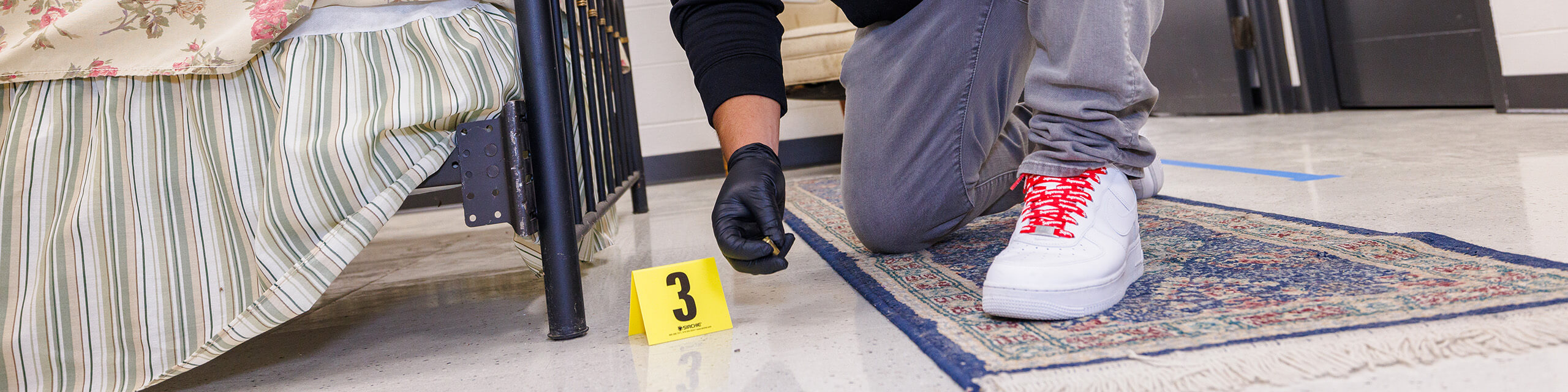 Person kneeling beside a bed with an evidence marker on the floor.